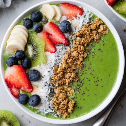 A vibrant green smoothie bowl topped with granola, sliced bananas, and fresh berries.